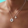 Two silver necklaces with circular pendants on a person's neck against a turquoise background